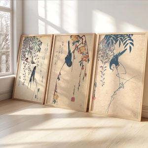 May include: Three framed prints with Japanese bird illustrations. Each print depicts a bird on a branch with flowers and foliage. The artwork uses muted tones of blue, red, and green against a beige background, creating a serene aesthetic.