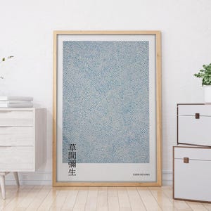May include: A framed print of a blue and white abstract art piece with the text "点描画 生" in black. The print is hanging on a white wall above a white dresser with two drawers. There is a small potted plant on a white chest of drawers to the right of the print.