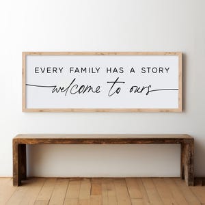 May include: A rectangular wooden framed sign with the text "EVERY FAMILY HAS A STORY welcome to ours" in black. The sign is mounted on a white wall above a dark brown wooden bench.