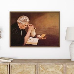 Op de afbeelding: Een ingelijste prent van een oudere persoon met wit haar, gekleed in het zwart, die bidt over een open boek. Een witte kan en brood staan op een tafel. Het kunstwerk heeft een warm, sepia-getinte kleurenpalet.