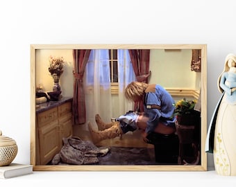 May include: A man wearing a blue shirt and brown boots sits on a toilet in a bathroom. The bathroom has a window with white curtains and a wooden cabinet with drawers.