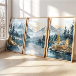 以下が含まれることがあります： 穏やかな山の湖の風景を描いた、額入りの風景画プリント3点。青、グレー、ゴールドの色調で水彩画風に描かれています。木製のフレームは白い壁に立てかけられており、穏やかな雰囲気を醸し出しています。
