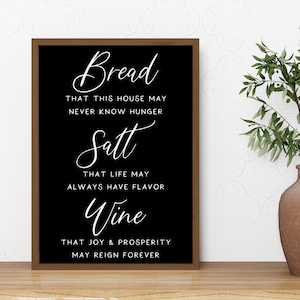 May include: Black framed sign with white text that reads "Bread that this house may never know hunger, Salt that life may always have flavor, Wine that joy & prosperity may reign forever."