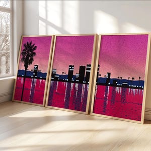 Peut inclure: Trois impressions encadrées avec une ville rose et violette avec un palmier au premier plan. Les impressions sont dans des cadres en bois et sont accrochées à un mur blanc.