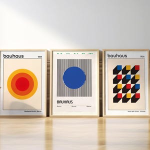 Könnte beinhalten: Drei gerahmte Drucke mit geometrischen Designs. Der erste Druck ist weiß mit einem roten, orangefarbenen und gelben Kreis in der Mitte. Der zweite Druck ist weiß mit einem blauen Kreis auf einem schwarz-weiß gestreiften Hintergrund. Der dritte Druck ist weiß mit einem schwarz-weißen Schachbrettmuster mit roten, gelben und blauen Quadraten. Alle drei Drucke haben das Wort "bauhaus" in schwarzer Schrift oben.
