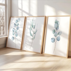 May include: Three framed botanical prints showcasing watercolour eucalyptus branches in shades of blue and green. The prints have a minimalist style with a white background and are displayed in light brown wooden frames.