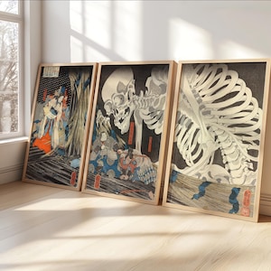 May include: Three framed Japanese woodblock prints leaning against a wall. The prints feature detailed illustrations of skeletons and figures in traditional attire, with a color palette of black, white, red, and blue. The frames are a light wood color.