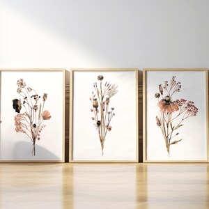 May include: Three framed prints of watercolour paintings of dried flowers in shades of brown, pink, and white. The prints are arranged in a row on a wooden floor.