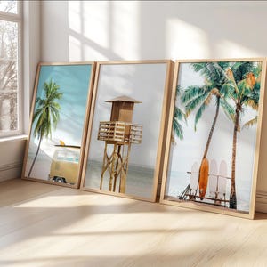 Puede incluir: Tres impresiones enmarcadas con temática de playa, apoyadas contra una pared. Las impresiones presentan una palmera, una torre de salvavidas y palmeras con tablas de surf. Los marcos son de madera clara y las imágenes tienen un ambiente soleado y costero.