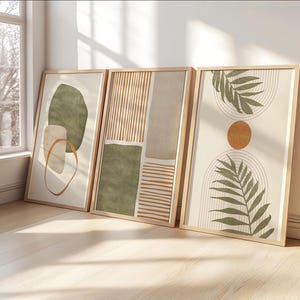 Op de afbeelding: Drie ingelijste abstracte kunstprints. De prints bevatten aardetinten van groen, beige en bruin. De kunst omvat geometrische vormen, lijnen en botanische elementen. De frames zijn gemaakt van lichtgekleurd hout.