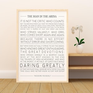 May include: Framed print with the text "The Man in the Arena" by Theodore Roosevelt. The quote is in black font on a white background, framed in a light wood frame. A small white vase with a plant is on a shelf.