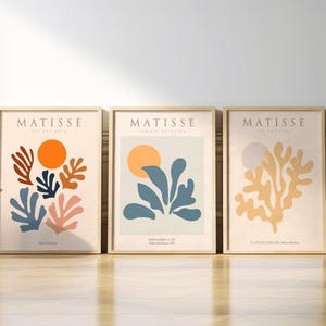 May include: Three framed art prints with a minimalist design featuring abstract shapes and colours. The prints are titled "Matisse" and feature a yellow sun, blue and orange leaves, and a yellow and orange leaf design.