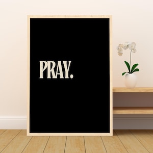 Op de afbeelding: Een ingelijste kunstdruk met een zwarte achtergrond en het woord "PRAY." in witte, vette, schreefletter. De lijst heeft een lichte houtkleur. De print wordt weergegeven op een houten oppervlak.