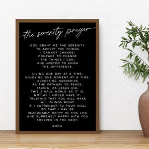 Puede incluir: Una impresión en blanco y negro enmarcada de "La Oración de la Serenidad" en una fuente manuscrita. La impresión está en un marco marrón y se muestra sobre una superficie de madera. Un jarrón con vegetación está a la derecha.