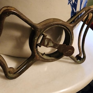 May include: A rusty metal clamp with a lever and a circular opening. The clamp is designed to hold a round object in place.