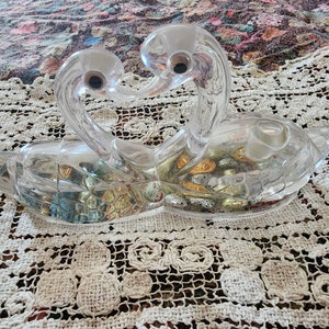 Puede incluir: Un par de figuras de cisnes de cristal transparente, enfrentadas con sus cuellos entrelazados. Los cisnes están sentados sobre un encaje de ganchillo blanco.