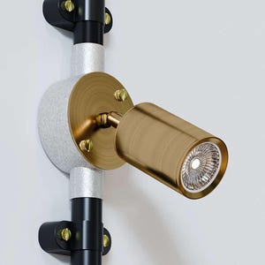 Puede incluir: Un foco moderno en tonos latón, montado en un sistema de riel negro y plateado. El foco tiene un cabezal cilíndrico con una luz empotrada brillante. El accesorio está fijado a una pared blanca.