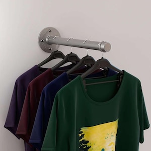 Puede incluir: Un perchero de pared de metal plateado con cuatro camisetas colgadas de perchas negras. Las camisetas son de color morado, burdeos, azul marino y verde. El perchero está fijado a la pared.