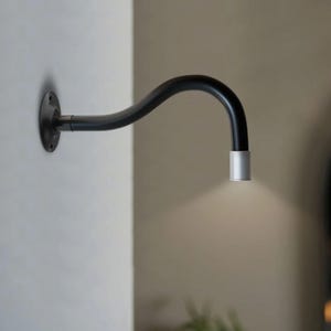 Puede incluir: Una lámpara de pared negra y curvada con un cabezal de luz plateado. La lámpara está fijada a una pared blanca con una placa de montaje circular. La luz emite un brillo suave.