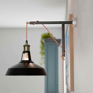 May include: A black metal wall lamp with a conical shade and a brass-coloured fixture. The lamp has a pivoting arm and a decorative, twisted copper-coloured cord. The lamp is mounted on a light-coloured wall.
