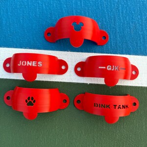 May include: A collection of five red, curved, plastic objects. One has a Mickey Mouse head cutout, another reads "JONES", a third reads "-GJK-", a fourth has a paw print, and the last reads "DINK TANK".