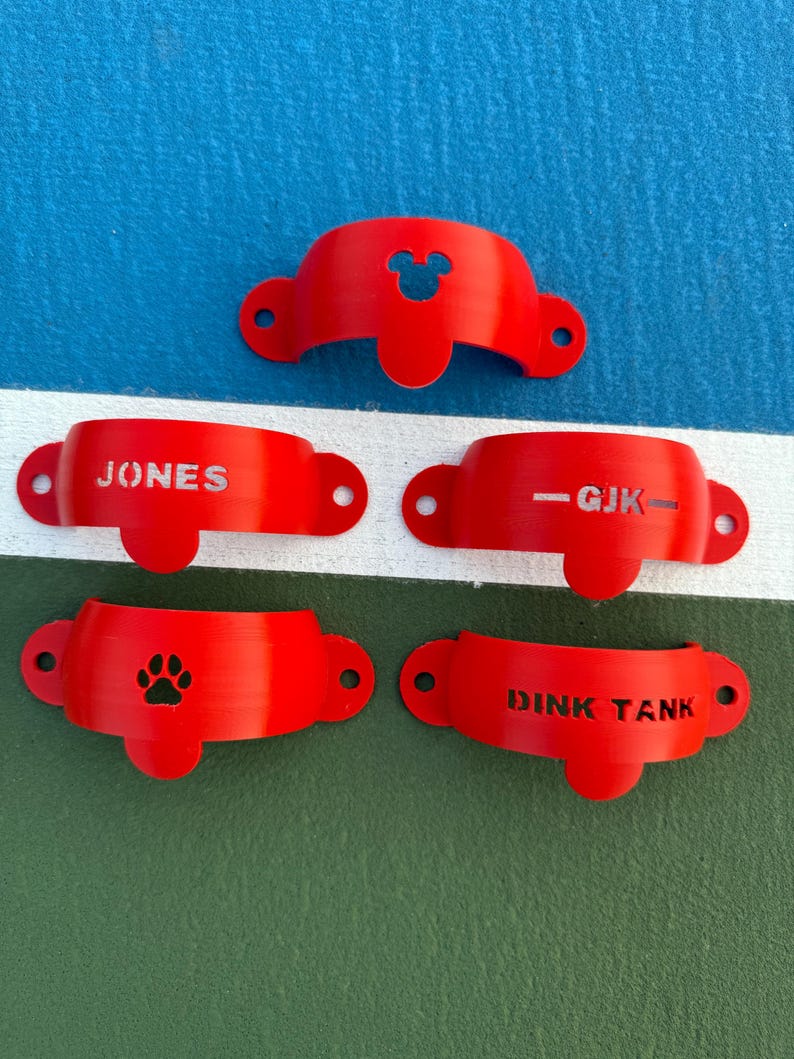 May include: A collection of five red, curved, plastic objects. One has a Mickey Mouse head cutout, another reads "JONES", a third reads "-GJK-", a fourth has a paw print, and the last reads "DINK TANK".