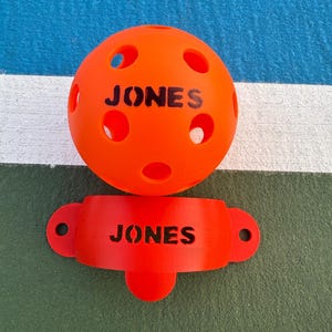 K&ouml;nnte beinhalten: Hellorangefarbener Pickleball-Ball und Paddelhalter. Der Ball ist rund mit mehreren L&ouml;chern und dem Wort "JONES" darauf gedruckt. Der Paddelhalter hat eine gebogene Form mit dem Wort "JONES" darauf.
