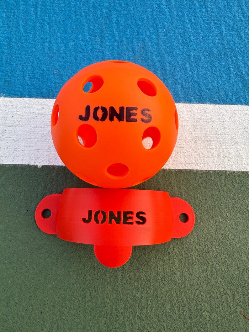 K&ouml;nnte beinhalten: Hellorangefarbener Pickleball-Ball und Paddelhalter. Der Ball ist rund mit mehreren L&ouml;chern und dem Wort "JONES" darauf gedruckt. Der Paddelhalter hat eine gebogene Form mit dem Wort "JONES" darauf.
