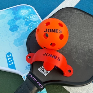 K&ouml;nnte beinhalten: Nahaufnahme von Pickleball-Ausr&uuml;stung. Ein orangefarbener Ball und ein roter Ballhalter, beide mit dem Namen "JONES", liegen auf einem schwarzen Schl&auml;ger. Ein hellblauer Schl&auml;ger ist ebenfalls sichtbar.