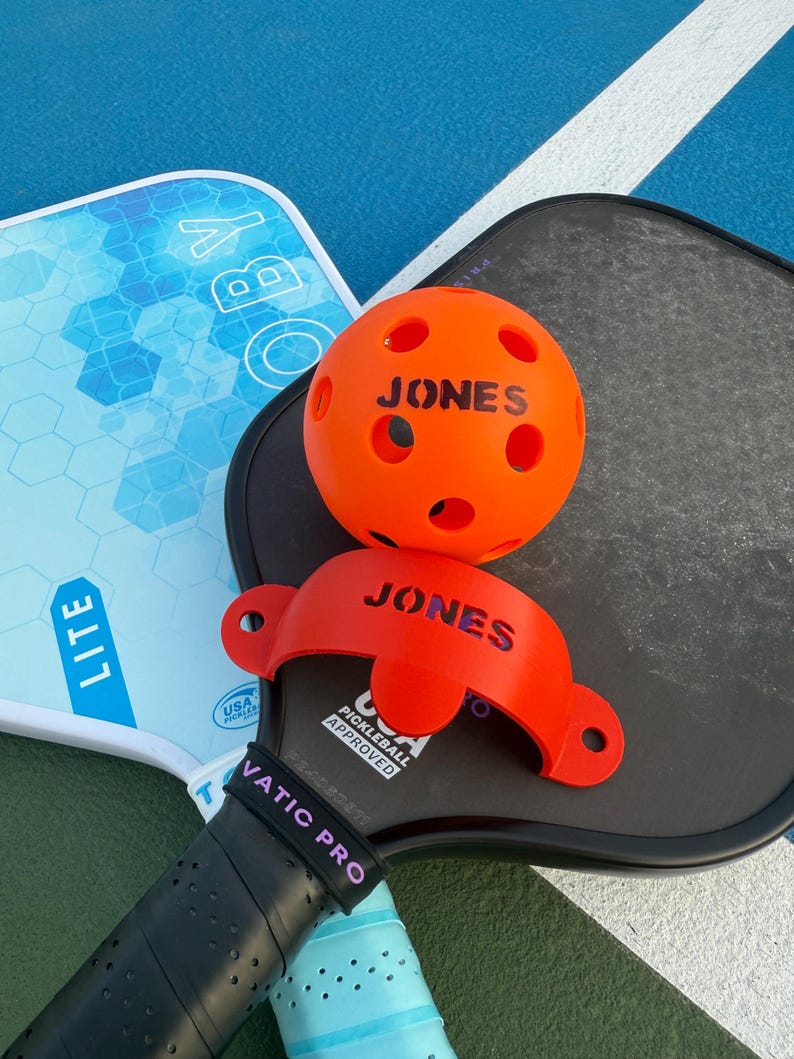 K&ouml;nnte beinhalten: Nahaufnahme von Pickleball-Ausr&uuml;stung. Ein orangefarbener Ball und ein roter Ballhalter, beide mit dem Namen "JONES", liegen auf einem schwarzen Schl&auml;ger. Ein hellblauer Schl&auml;ger ist ebenfalls sichtbar.
