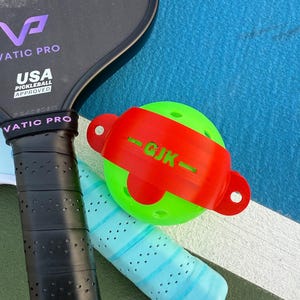 May include: A close-up of pickleball equipment, including a black paddle with the text "VATIC PRO" and "USA PICKLEBALL APPROVED." A green pickleball is secured by a red band with the letters "GJK." A blue and black grip is also visible.
