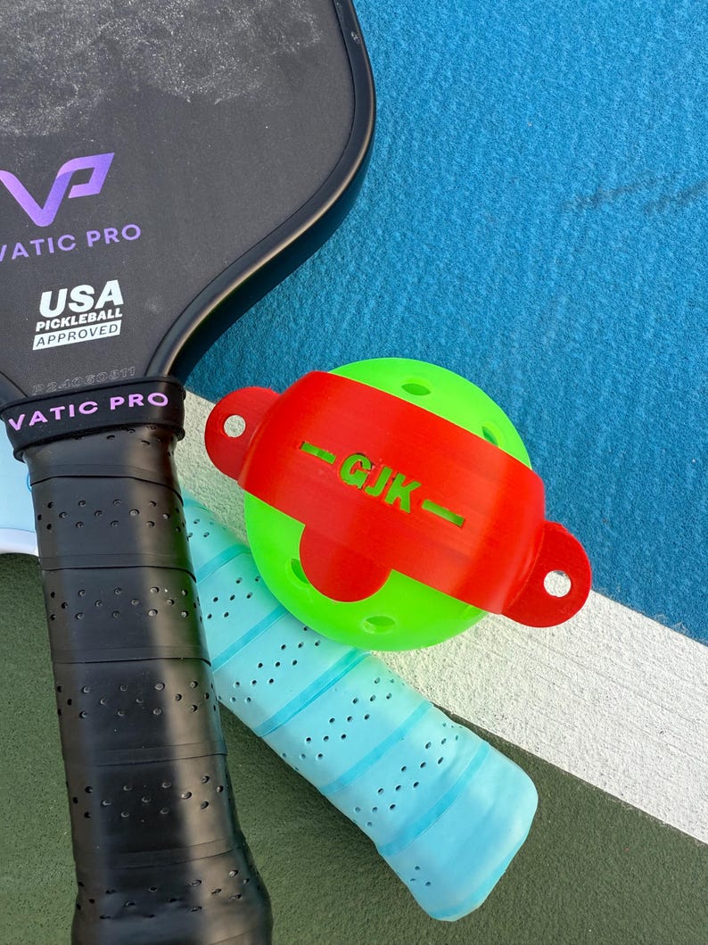 May include: A close-up of pickleball equipment, including a black paddle with the text "VATIC PRO" and "USA PICKLEBALL APPROVED." A green pickleball is secured by a red band with the letters "GJK." A blue and black grip is also visible.