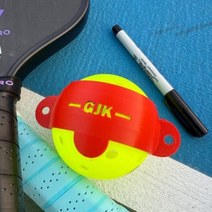 May include: A yellow pickleball with a red band marked "GJK" and a black Sharpie marker on a blue surface. A black pickleball paddle and a blue grip are also visible.