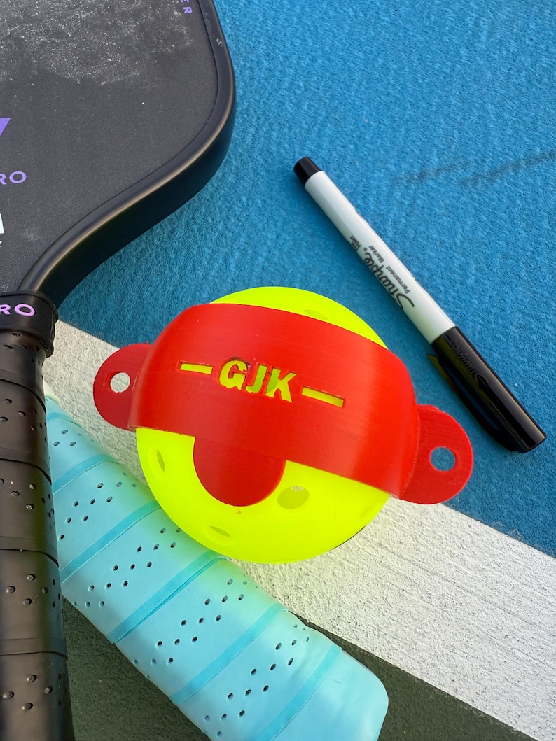 May include: A yellow pickleball with a red band marked "GJK" and a black Sharpie marker on a blue surface. A black pickleball paddle and a blue grip are also visible.