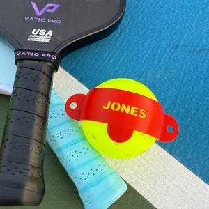 K&ouml;nnte beinhalten: Ein Pickleball-Set mit einem schwarzen Vatic Pro Schl&auml;ger, einem hellblauen Griff und einem gelben Ball mit einem roten Band mit der Aufschrift "JONES". Der Schl&auml;ger ist von USA Pickleball zugelassen.