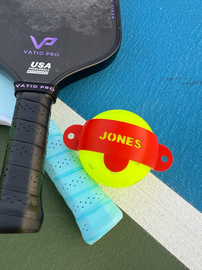 K&ouml;nnte beinhalten: Ein Pickleball-Set mit einem schwarzen Vatic Pro Schl&auml;ger, einem hellblauen Griff und einem gelben Ball mit einem roten Band mit der Aufschrift "JONES". Der Schl&auml;ger ist von USA Pickleball zugelassen.