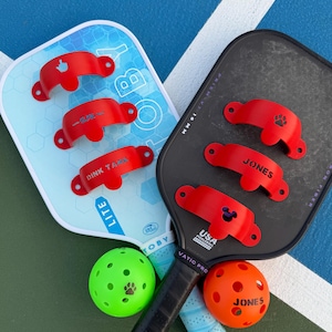 K&ouml;nnte beinhalten: Zwei Pickleball-Schl&auml;ger, einer hellblau und einer schwarz, mit roten Accessoires. Auf dem hellblauen Schl&auml;ger stehen die W&ouml;rter "TOBY" und "LITE". Zwei Pickleballs, einer gr&uuml;n und einer orange, sind ebenfalls zu sehen.