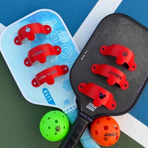 May include: Two pickleball paddles, one light blue and one black, with red accessories. The light blue paddle has the word "TOBY" and "LITE" printed on it. Two pickleballs, one green and one orange, are also in the image.