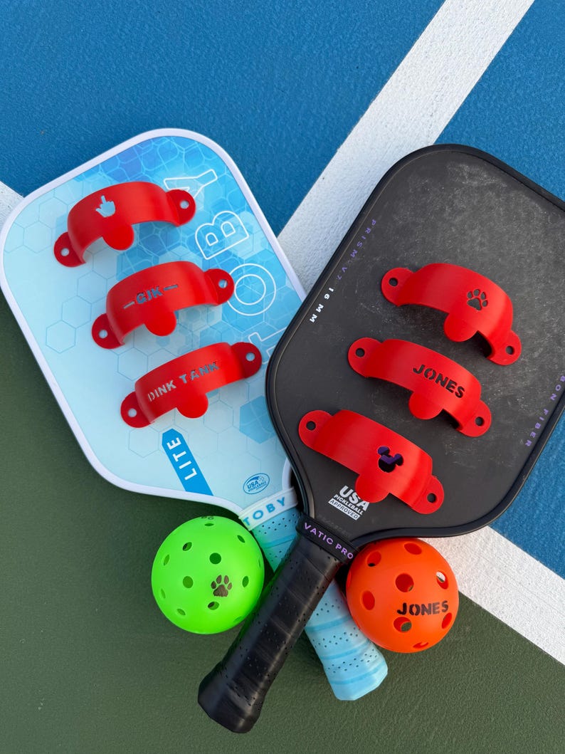 May include: Two pickleball paddles, one light blue and one black, with red accessories. The light blue paddle has the word "TOBY" and "LITE" printed on it. Two pickleballs, one green and one orange, are also in the image.