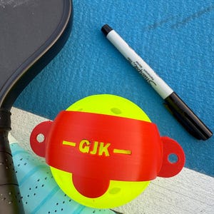 May include: A yellow pickleball with a red band featuring the initials "GJK" and a black Sharpie marker on a blue surface. A black pickleball paddle and a blue and white grip are also visible.