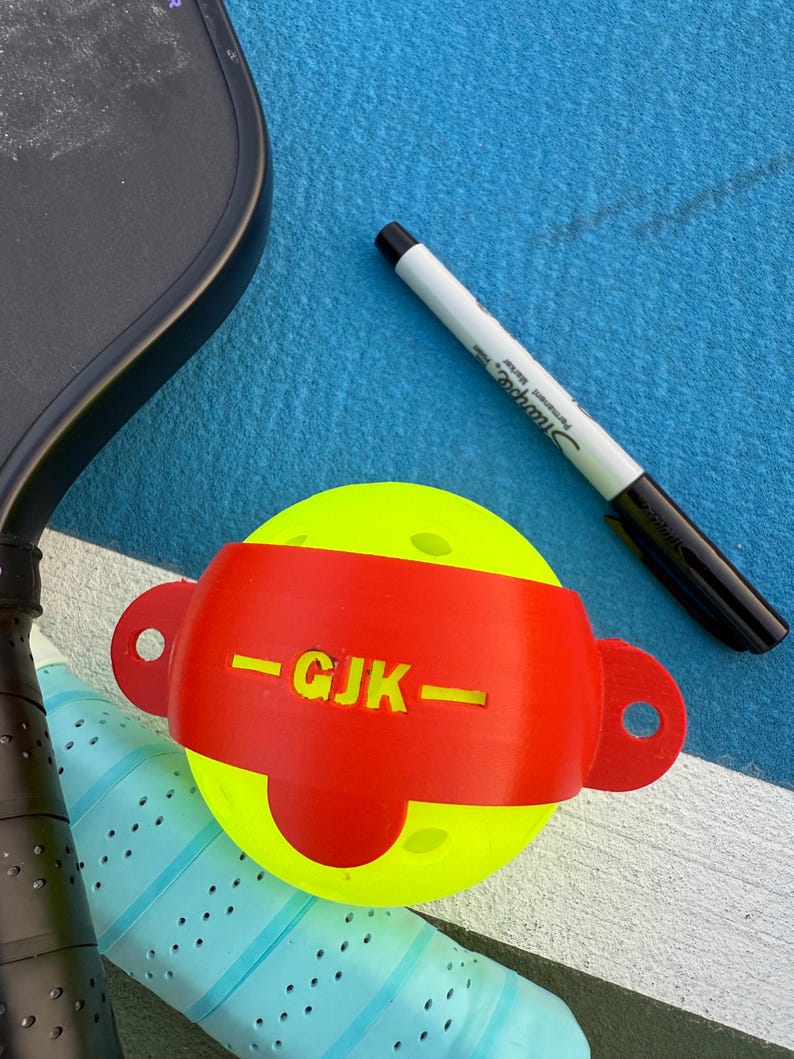 May include: A yellow pickleball with a red band featuring the initials "GJK" and a black Sharpie marker on a blue surface. A black pickleball paddle and a blue and white grip are also visible.