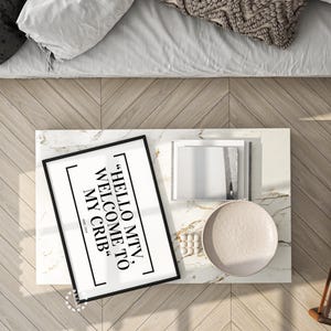 Könnte beinhalten: Draufsicht auf einen Couchtisch mit Marmorplatte und einem gerahmten Druck mit der Aufschrift "HELLO MTV, WELCOME TO MY CRIB". Auf dem Tisch befinden sich auch ein Stapel Bücher, ein Teller und ein kleines dekoratives Element. Der Tisch steht neben einem Bett mit weißer Bettwäsche.