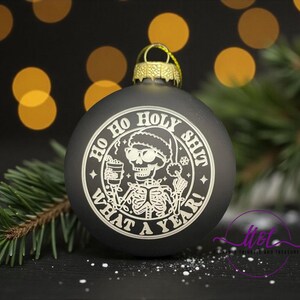 May include: Black Christmas ornament with a gold cap and a white graphic of a skeleton wearing a Santa hat, holding a drink, and the text "Ho Ho Holy Shit What A Year!" Festive holiday decor.