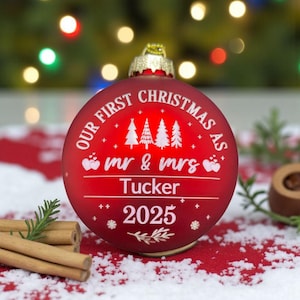 May include: A red Christmas ornament with the text "OUR FIRST CHRISTMAS AS" and "mr & mrs Tucker 2025" in white. The ornament features a gold cap and is decorated with Christmas trees and small star-like shapes. Cinnamon sticks and a sprig of greenery are in the foreground.