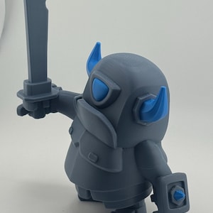 May include: A gray figurine of a stylized armored character with blue horns, a blue visor, and a blue accent on the shield. The character holds a gray sword in its right hand and wears a chest plate, standing in a dynamic pose with a rounded body.