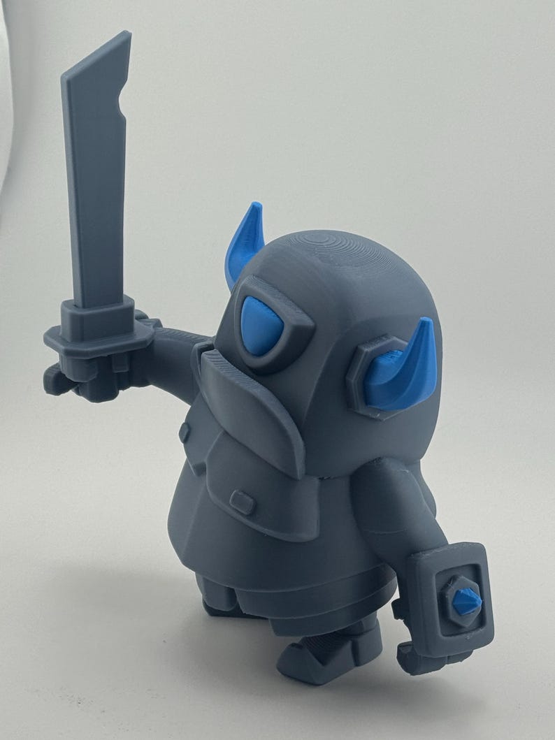 May include: A gray figurine of a stylized armored character with blue horns, a blue visor, and a blue accent on the shield. The character holds a gray sword in its right hand and wears a chest plate, standing in a dynamic pose with a rounded body.