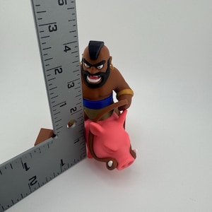 Puede incluir: Una figura de Clash of Clans con un personaje de piel morena con un mohawk negro, barba y pendientes dorados, sosteniendo un cerdo rosa. La figura mide aproximadamente 10 cm de altura.