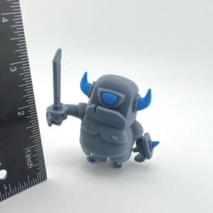 May include: A gray figurine with blue horns and a blue eye, holding a sword in its right hand. A black ruler is positioned to the left, showing the figure stands approximately 2.5 inches tall. The character has a blocky, armored appearance.