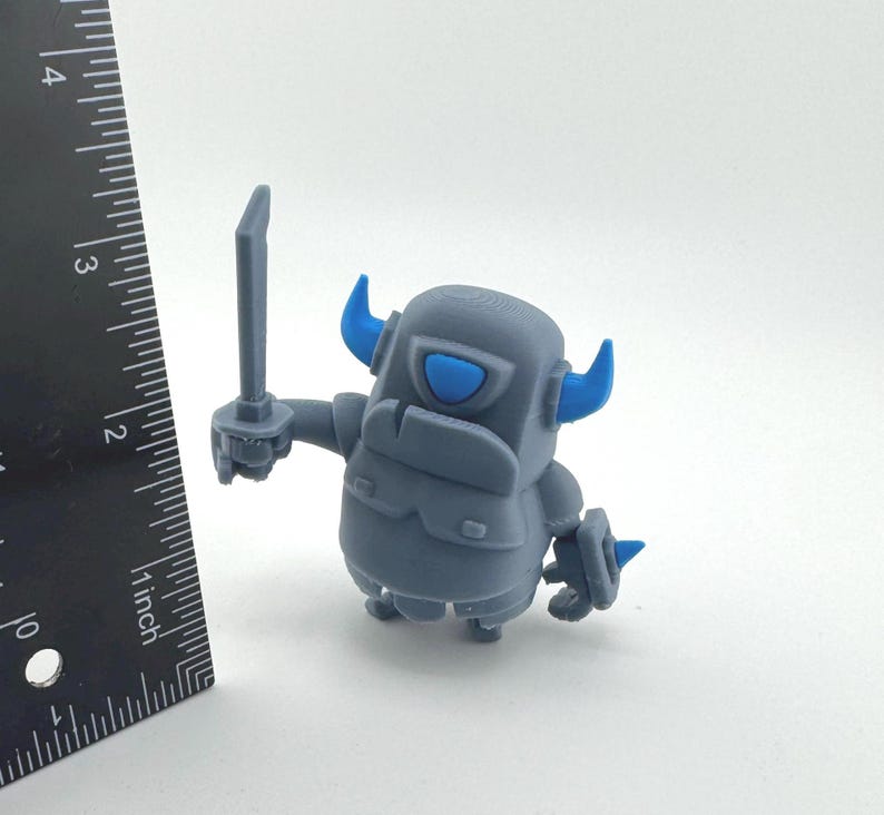 May include: A gray figurine with blue horns and a blue eye, holding a sword in its right hand. A black ruler is positioned to the left, showing the figure stands approximately 2.5 inches tall. The character has a blocky, armored appearance.