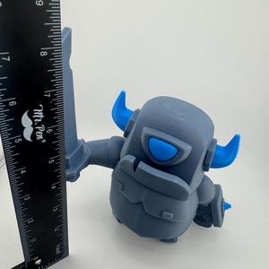 May include: A gray and blue plastic figurine of a knight character holding a sword, positioned next to a black Mr. Pen ruler. The ruler shows the figurine stands approximately 5.5 inches tall from the base to the top of the sword blade.